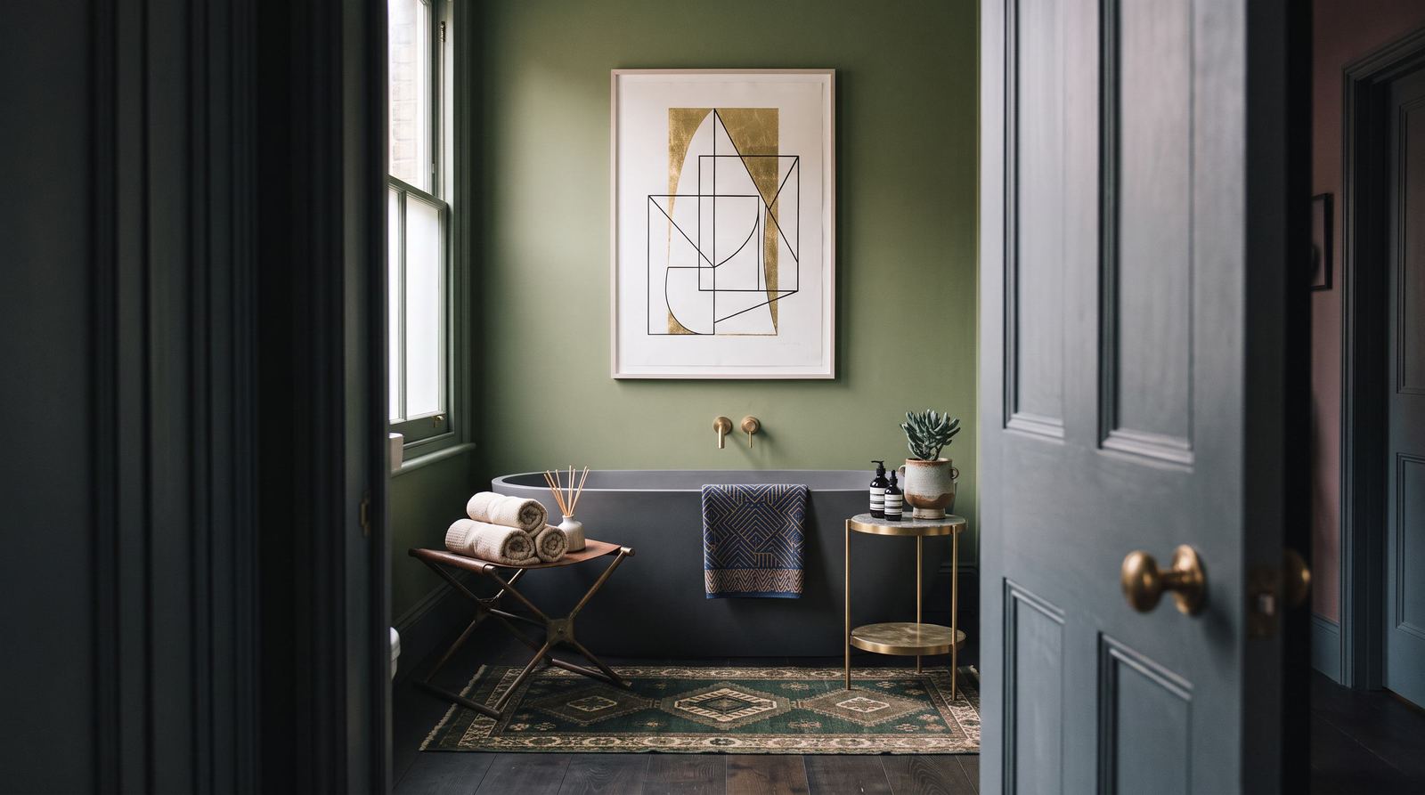 A sage-green bathroom with a freestanding grey bath and framed artwork, seen through an open panelled door